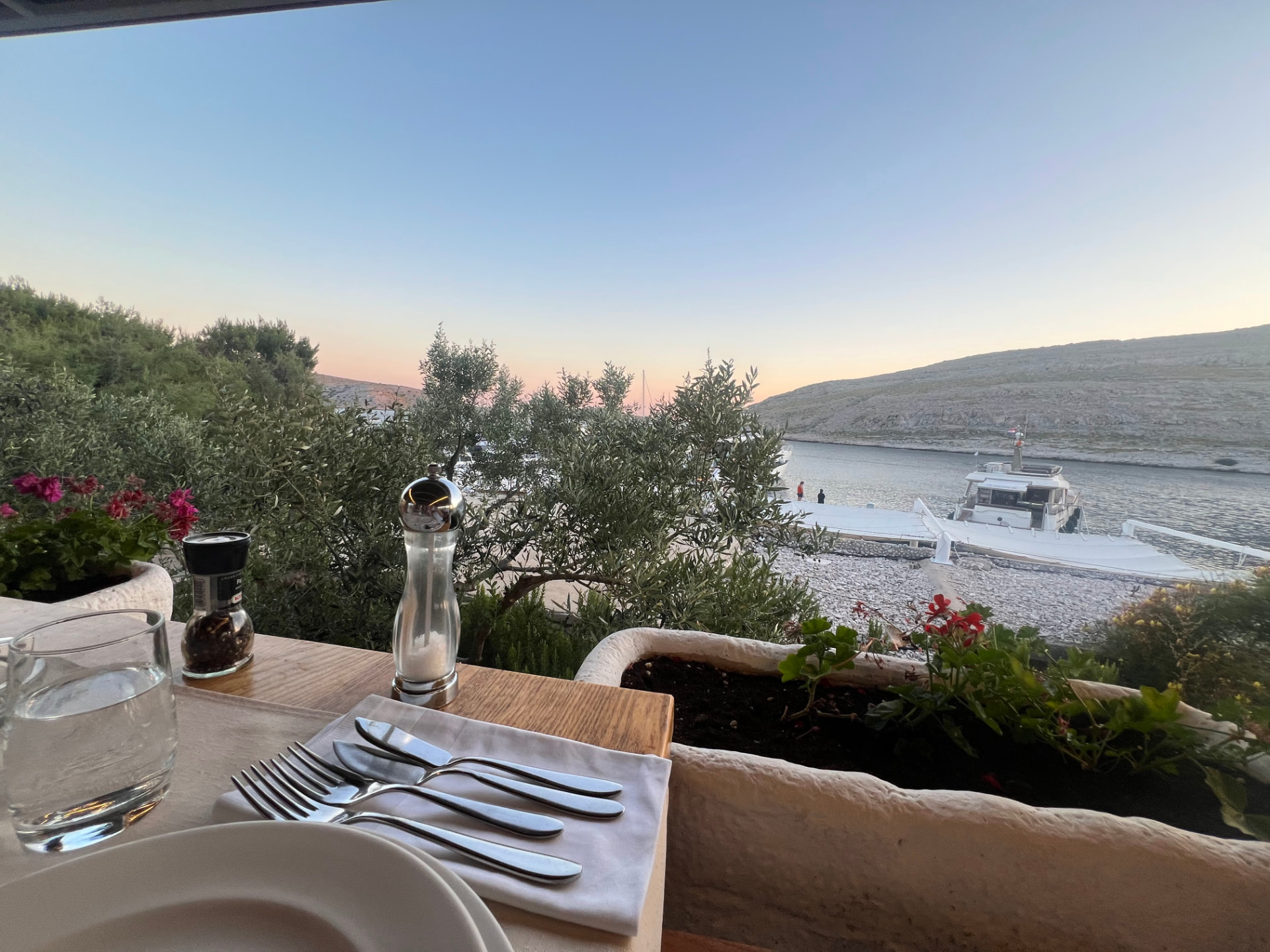 A laid table above the cove at dusk with olive trees and a moored boat